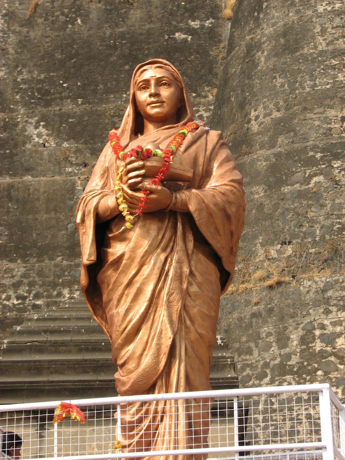 Bronze statue of Ahilyabai Holkar wearing a garland of flowers, standing before a stone fort wall above a flight of steps.